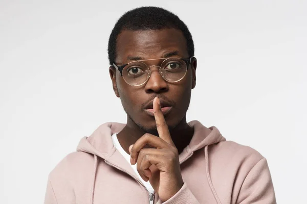 Imagen de estudio del joven guapo africano aislado sobre fondo blanco con anteojos redondos y presionando el dedo índice a los labios como si pidiese a otros que guarden silencio sobre el emocionante secreto o los datos —  Fotos de Stock