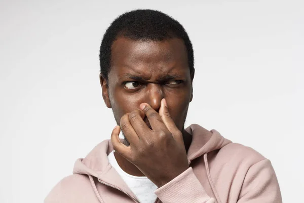 Indoor portret van knappe African American guy afgebeeld geïsoleerd op witte achtergrond houden zijn neus probeert te ontsnappen aan stinkende geur en opzij kijken met zorg op zoek naar de bron van de geur — Stockfoto