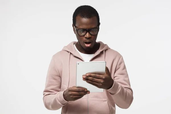 Horizontale Nahaufnahme eines jungen afrikanisch-amerikanischen Mannes, der isoliert auf weißem Hintergrund in lässiger Kleidung und Brille abgebildet ist und einen erstaunten Blick mit offenem Mund auf den Tablet-Bildschirm wirft, den er in der Hand hält — Stockfoto