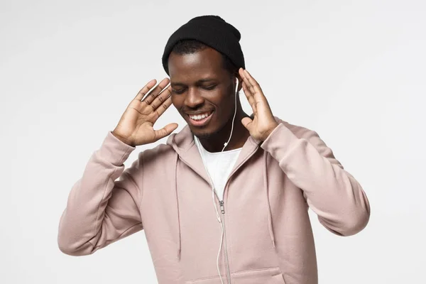 Binnen foto van knappe Afro-Amerikaanse jongen geïsoleerd op een witte achtergrond, het dragen van muts en hoodie oprijzen handen terwijl het genieten van audio-track door witte koptelefoon, glimlachend met ogen gesloten — Stockfoto