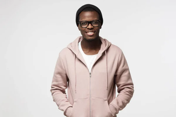 Horizontale Aufnahme eines jungen positiv aussehenden afrikanischen Männchens isoliert auf weißem Hintergrund stehend in ruhiger entspannter Haltung in rosa Kapuzenpulli, schwarzem Hut und Brille, die selbstbewusst lächeln — Stockfoto