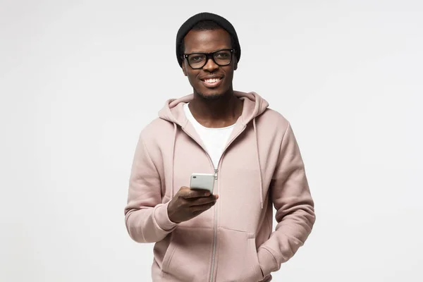 Nahaufnahme Porträt eines jungen afrikanisch-amerikanischen Mannes in lässiger Kleidung und Brille, der isoliert auf weißem Hintergrund abgebildet ist und sein Handy in der Hand hält und mit offenem Lächeln in die Kamera schaut, surft oder SMS schreibt — Stockfoto