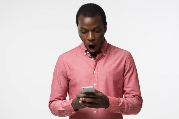 Horizontales Porträt eines gut aussehenden afrikanisch-amerikanischen Kerls, der isoliert auf weißem Hintergrund abgebildet ist und auf den Bildschirm des Smartphones mit rundem Mund schaut, weil er tief erstaunt ist und große Neuigkeiten verkündet. — Stockfoto
