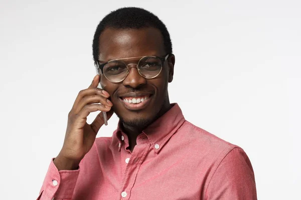 Nahaufnahme Bild eines jungen afrikanischen Hipster-Mannes isoliert auf weißem Hintergrund mit aufgesetzter runder Brille, der das Telefon ans Ohr drückt, während er mit einem Kollegen oder Freund spricht, lächelnd — Stockfoto