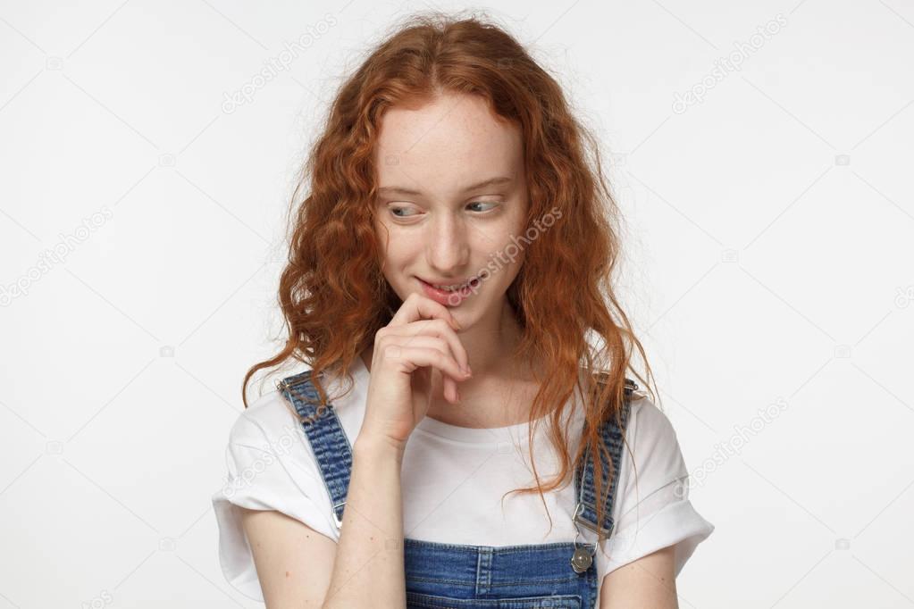 Closeup headshot of young attractive European woman with red loose curly hair isolated on white background touching her chin and looking down while considering new opportunities with timid smile