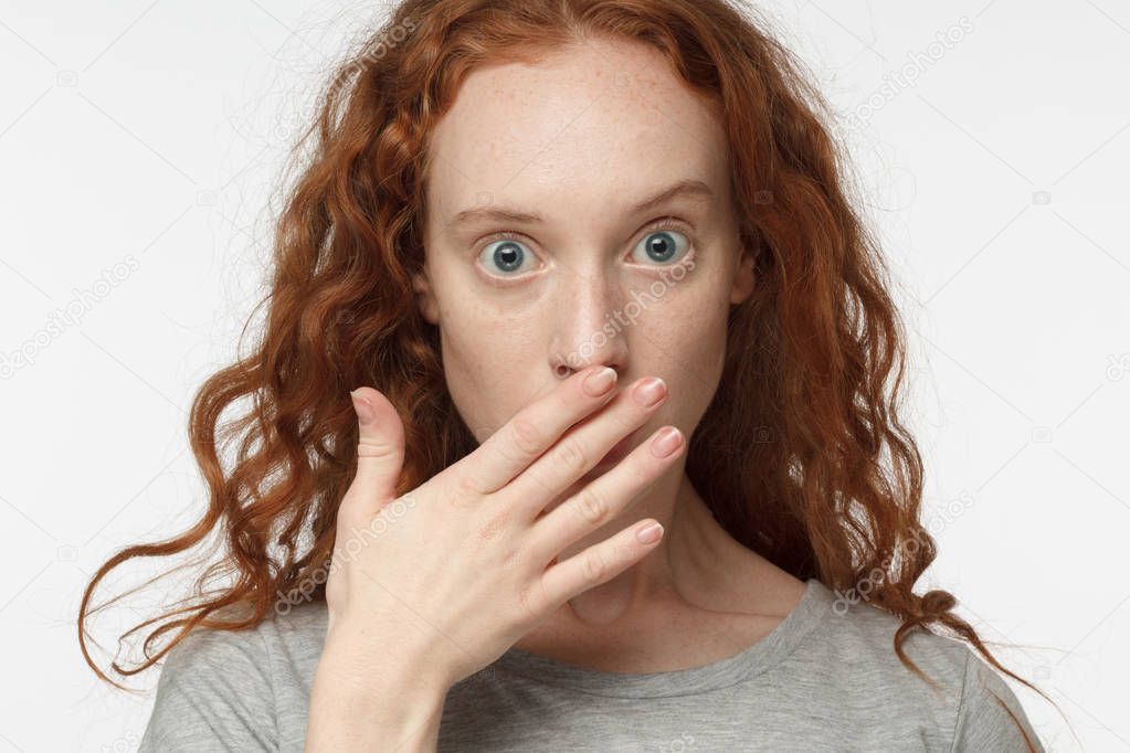 Astonished bug-eyed curly redhead girl covering her mouth with one hand, looking shocked. Surprised, embarrassed and confused female showing omg emotion