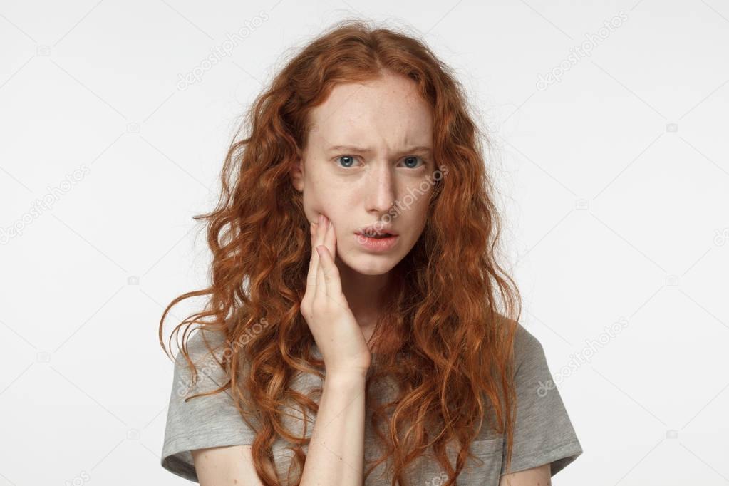 Indoor portrait of redhead girl feeling pain, holding her cheek with hand, suffering from bad toothache, looking at camera with painful expression. Tooth ache concept.