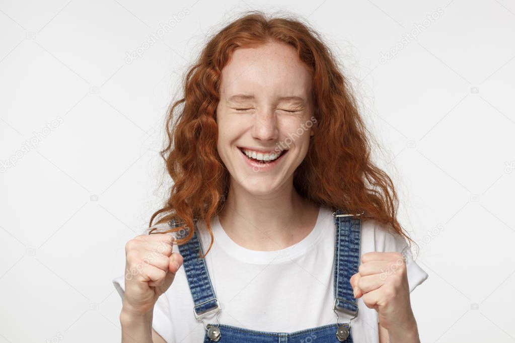 Closeup of emotional redhead girl isolated on gray background showing white teeth while screaming with joy and victorious expression, holding hands in gesture of winner, looking extremely happy