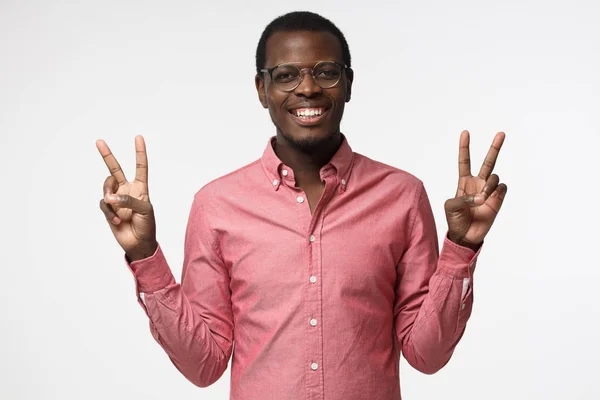 Indoor portret van jonge Afrikaanse man geïsoleerd op grijze achtergrond in casual kleding met optimistisch glimlach, tonen overwinning teken met beide handen, op zoek vriendelijk en bereid om te verwelkomen — Stockfoto