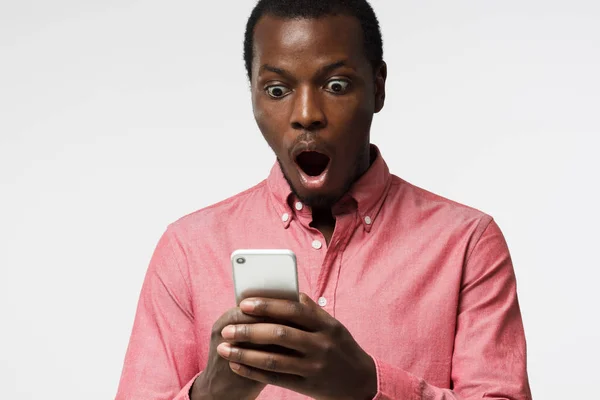 Nahaufnahme Porträt eines hübschen jungen Schwarzen in rosa Hemd, Smartphone in der Hand, erschrocken und verwirrt beim Lesen von SMS oder E-Mail — Stockfoto