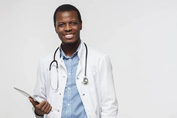 Foto horizontal del médico africano aislado sobre fondo gris sonriendo felizmente y sosteniendo su tablet. Concepto de aparatos y servicios médicos contemporáneos — Foto de Stock