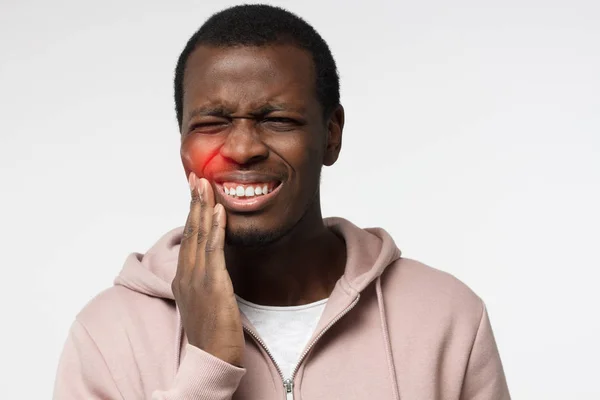 Retrato interior de un joven afroamericano preocupado aislado sobre fondo blanco vestido con camiseta blanca y sudadera con capucha rosa que experimenta un fuerte dolor de muelas presionando la mano contra la barbilla debido al dolor de mandíbulas —  Fotos de Stock