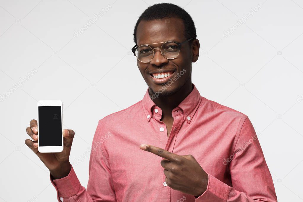 Happy african male isolated on grey background demonstrating blank smartphone and pointing to screen with expression of joy and interest. Copyspace for advertising products and services.