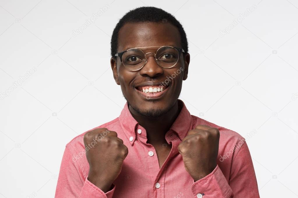 Closeup of emotional african male isolated on gray background showing white teeth, smiling with joy and victorious expression, holding hands in gesture of winner, looking extremely happy