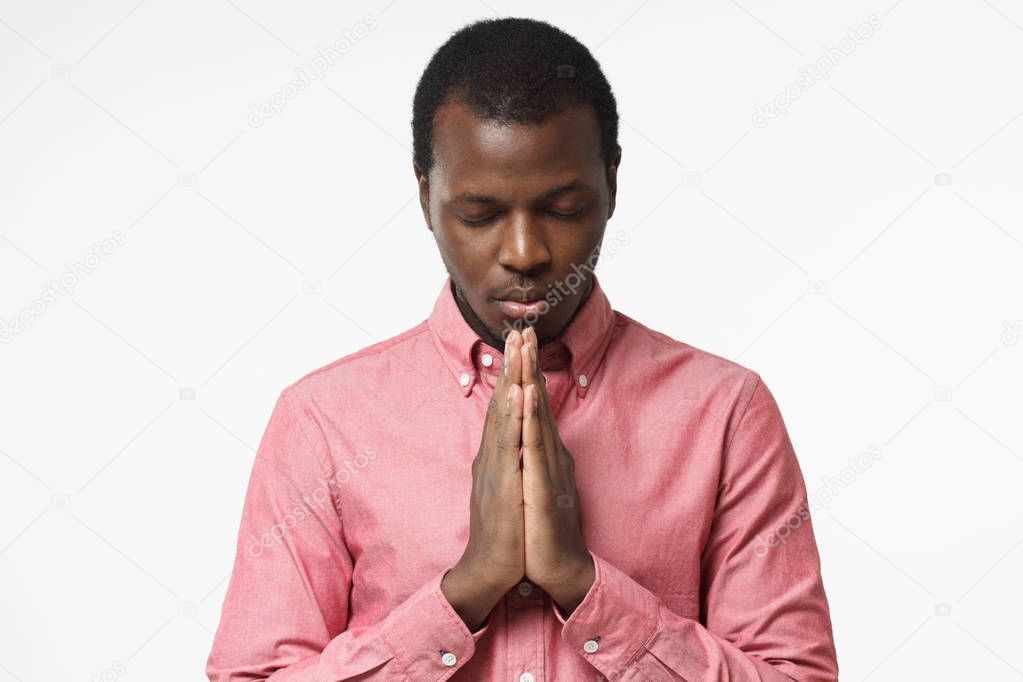 Closeup of young african american man isolated on gray background looking stressed, putting hands together as if he is praying with closed eyes to overcome depression
