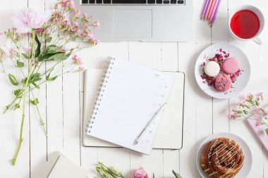Kadınsı düz çalışma alanı laptop, fincan çay, planlayıcısı, macarons ve beyaz tahta masaya çiçek ile yatıyordu. Üstten Görünüm kadar sahte.