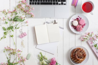 Kadınsı düz çalışma alanı laptop, fincan çay, planlayıcısı, macarons ve beyaz tahta masaya çiçek ile yatıyordu. Üstten Görünüm kadar sahte.