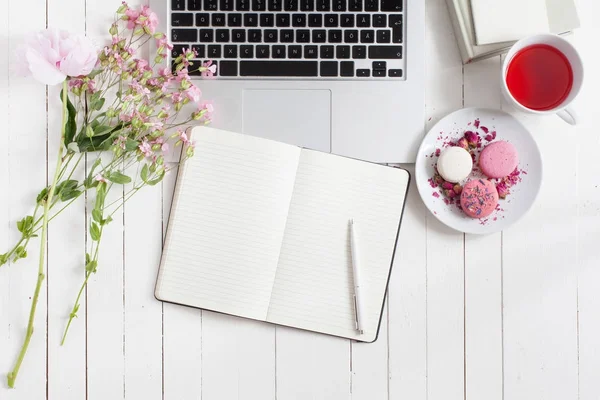 Espace de travail féminin plat avec ordinateur portable, tasse de thé, macarons et fleurs, planificateur et stylo sur table en bois blanc. Vue du dessus maquette . — Photo