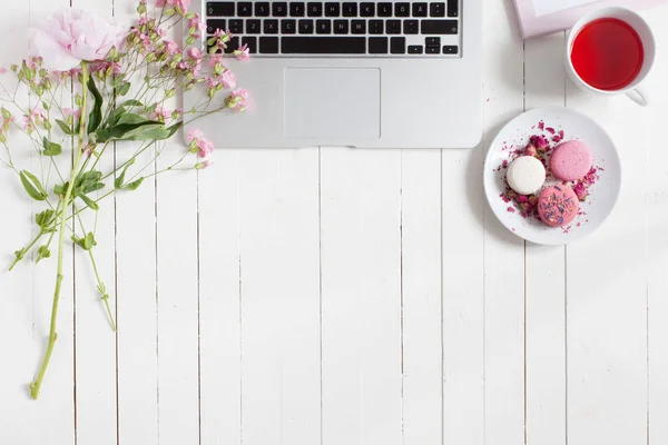 Жіночий плоский робочий простір з ноутбуком, чашкою чаю, макаронами та квітами на білому дерев'яному столі. Вид зверху сміється . — стокове фото