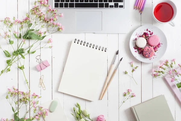 Espacio de trabajo femenino plano con portátil, taza de té, macarrones, flores, cuaderno, pinceles, sobre mesa de madera blanca. Vista superior maqueta hacia arriba . — Foto de Stock