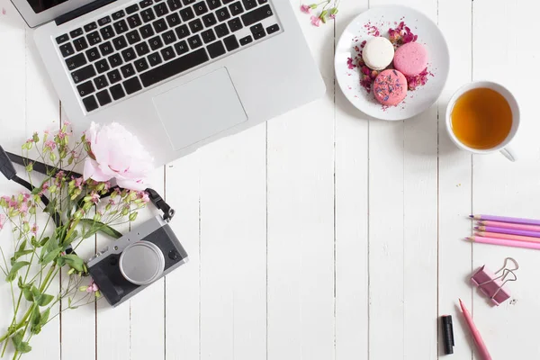 Ruang kerja datar feminin dengan laptop, secangkir teh, kamera retro, makaron dan bunga di meja kayu putih. tiruan top. Kosongkan ruang bagi teks Anda . — Stok Foto