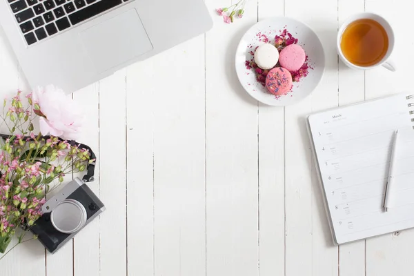 Ruang kerja datar feminin dengan laptop, secangkir teh, kamera retro, makaron dan bunga di meja kayu putih. tiruan top. Kosongkan ruang bagi teks Anda . — Stok Foto