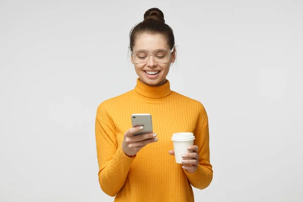 Inomhus bild av ung snygg Europeiska kvinna isolerade på grå bakgrund håller telefonen i handen, ser att skärmen leende medan du surfar eller läser meddelande — Stockfoto