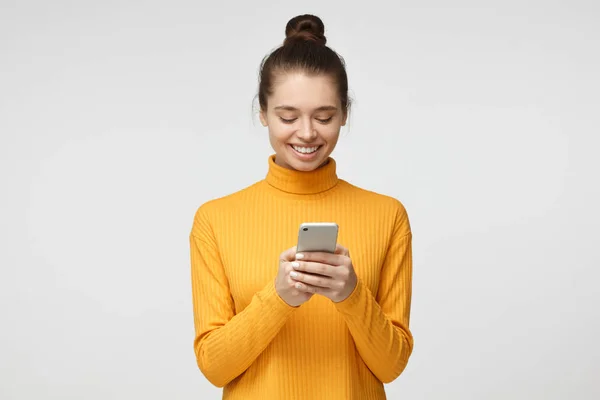 Yong vrij vrouwelijke staande geïsoleerd op grijze achtergrond aandachtig kijken naar scherm van cellphone, browsen van web-pagina's en mooi glimlachen tijdens het chatten — Stockfoto