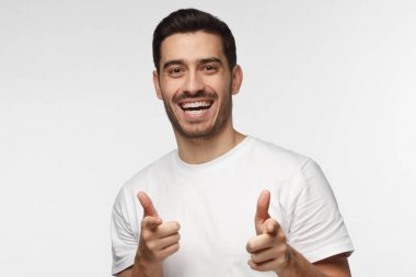 Hey siz! Genç adam kameraya gri arka plan üzerinde izole parmaklarıyla işaret