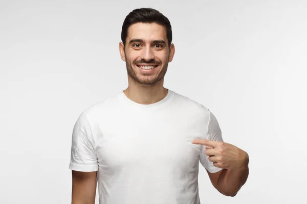 Young man pointing with index finger at blank white t-shirt with empty space for your advertising text or image, standing isolated on gray background — Stock Photo, Image