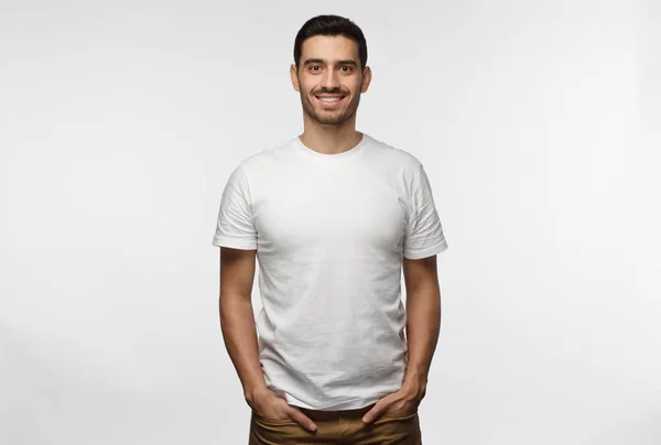 Young european man standing with hands in pockets, wearing blank white tshirt with copy space for your logo or text, isolated on grey background — Stock Photo, Image