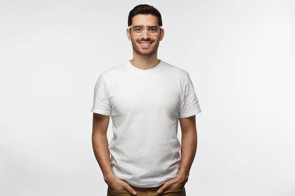 Handsome man in white tshirt and trasparent eyeglasses isolated on grey background, smiling, standing in hands in pockets pose — Stock Photo, Image