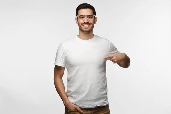 Jovem atraente em óculos transparentes apontando para sua camiseta branca em branco com o dedo indicador, mostrando espaço vazio para o seu texto publicitário ou logotipo, statning isolado em fundo cinza — Fotografia de Stock