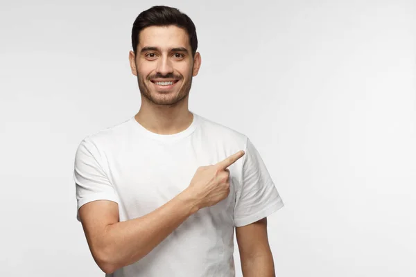 Jovem atraente em camiseta branca apontando para a direita com o dedo isolado em fundo cinza — Fotografia de Stock