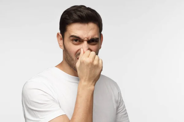 Portrait of young handsome man in casual clothes isolated on grey background holding his nose as if smelling something rotten and stinky looking aside trying to find source of odor — Stock Photo, Image