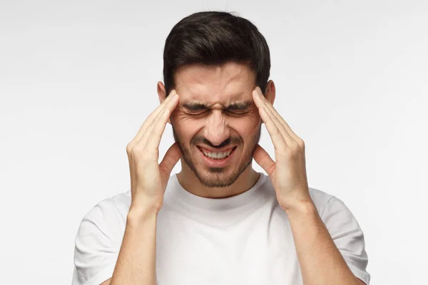 Portret van jonge man geïsoleerd op een grijze achtergrond die lijden aan ernstige hoofdpijn, drukken vingers aan tempels, sluiten de ogen voor het verlichten van pijn met hulpeloos gezicht expressie — Stockfoto