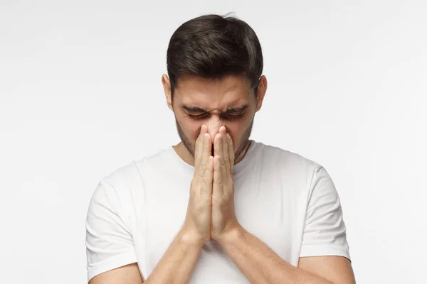 Body language. Sick young man in white t-shirt covering face with hands, sneezing, isolated on gray background — Stock Photo, Image