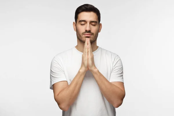 Closeup of young man in white tshirt isolated on gray background looking stressed, putting hands together as if he is praying with closed eyes to overcome depression — Stock Photo, Image