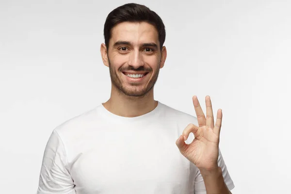Молодой человек в белой футболке, счастливый взгляд, улыбающийся, жестикулирующий, показывающий знак ОК. Белый мужчина показывает пальцами жест ОК. Концепция языка тела — стоковое фото