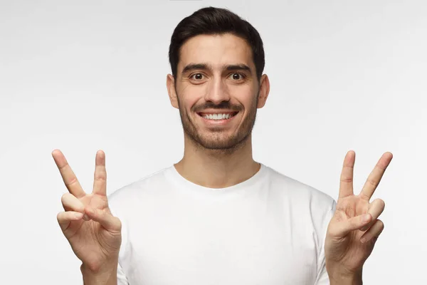 Крытый портрет молодого мужчины на сером фоне в белой футболке с оптимистической улыбкой, показывающий знак победы обеими руками, выглядящий дружелюбно и желающий приветствовать и общаться — стоковое фото