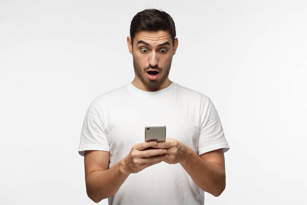 Close up portrait of handsome young man in white t-shirt, holding smartphone, looking scared and confused reading text message or e-mail