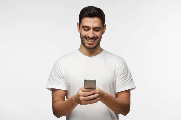 Крупный план человека в белой футболке, стоящего изолированно на сером фоне, внимательно смотрящего на экран мобильного телефона, просматривающего веб-страницы и красиво улыбающегося во время общения с друзьями — стоковое фото