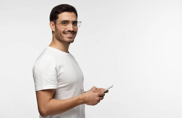 Jovem atraente homem de t-shirt branca olhando e sorrindo para a câmera com expressão alegre ao enviar mensagens de texto a seus amigos, usando telefone celular, isolado em fundo cinza — Fotografia de Stock