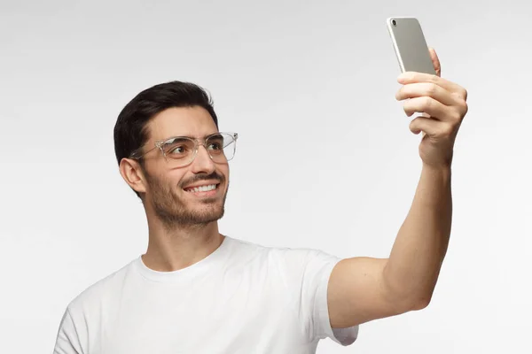 Портрет молодого красивого кавказца в белом тширте, стоящего на сером фоне с помощью смартфона, чтобы сделать селфи и улыбающийся — стоковое фото