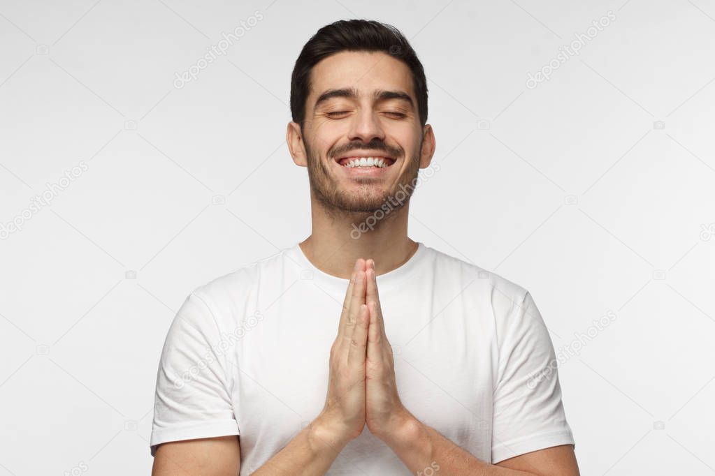 Young man isolated on grey background, having put hands together in prayer or meditation, smiling, dreaming and waiting for all best on exam