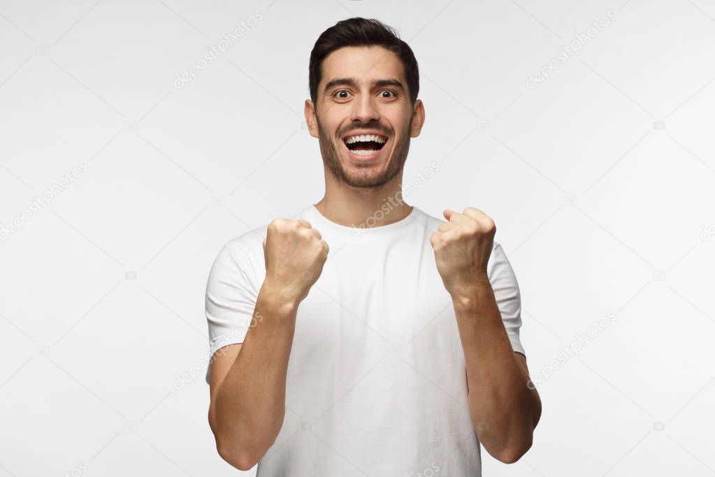 Indoor photo of good-looking man isolated on gray background celebrating victory and acting as if he is winner, squeezing fists in deep emotional expression of happiness and luck