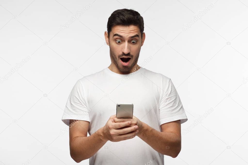 Portrait of young man isolated on gray background, looking agitated at display of her smartphone, impressed by media content from web