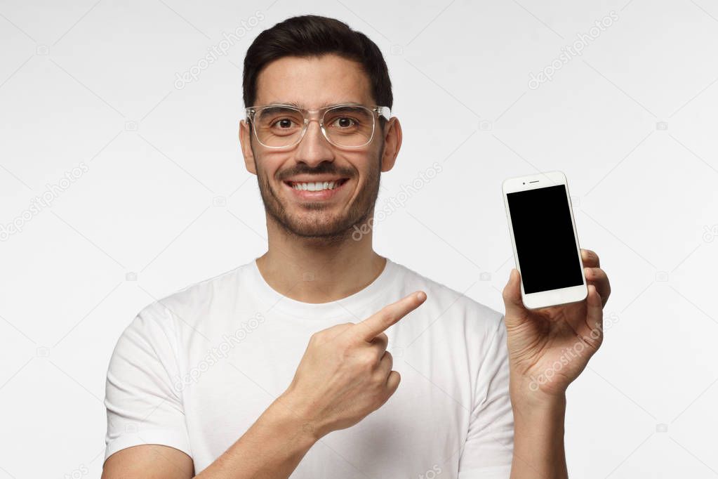 Handsome man in white tshirt in transparent glasses isolated on grey background, presenting smart phone and pointing with finger at blank black screen with copyspace for ads