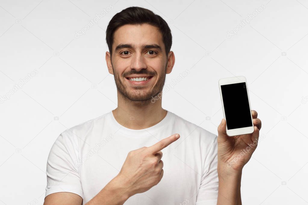 Indoor portrait of attractive young man isolated on grey background, holding blank smartphone, smiling at camera, feeling happy
