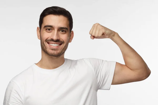 Alegre hombre atlético sonriente en camiseta blanca flexionando bíceps y mirando a la cámara aislada en el fondo gris — Foto de Stock